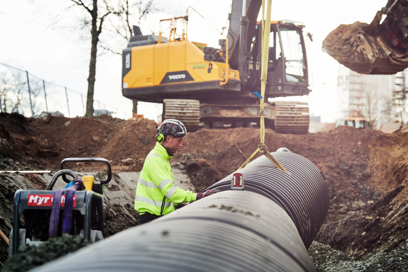 VA-arbete pågår med en man i varselkläder ståendes vid en VA-ledning. I bakgrunden syns en grävskopa.