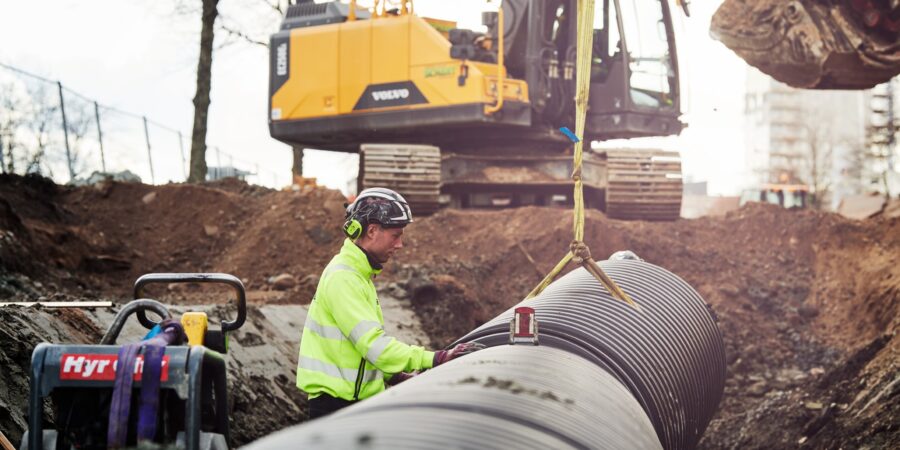 VA-arbete pågår med en man i varselkläder ståendes vid en VA-ledning. I bakgrunden syns en grävskopa.