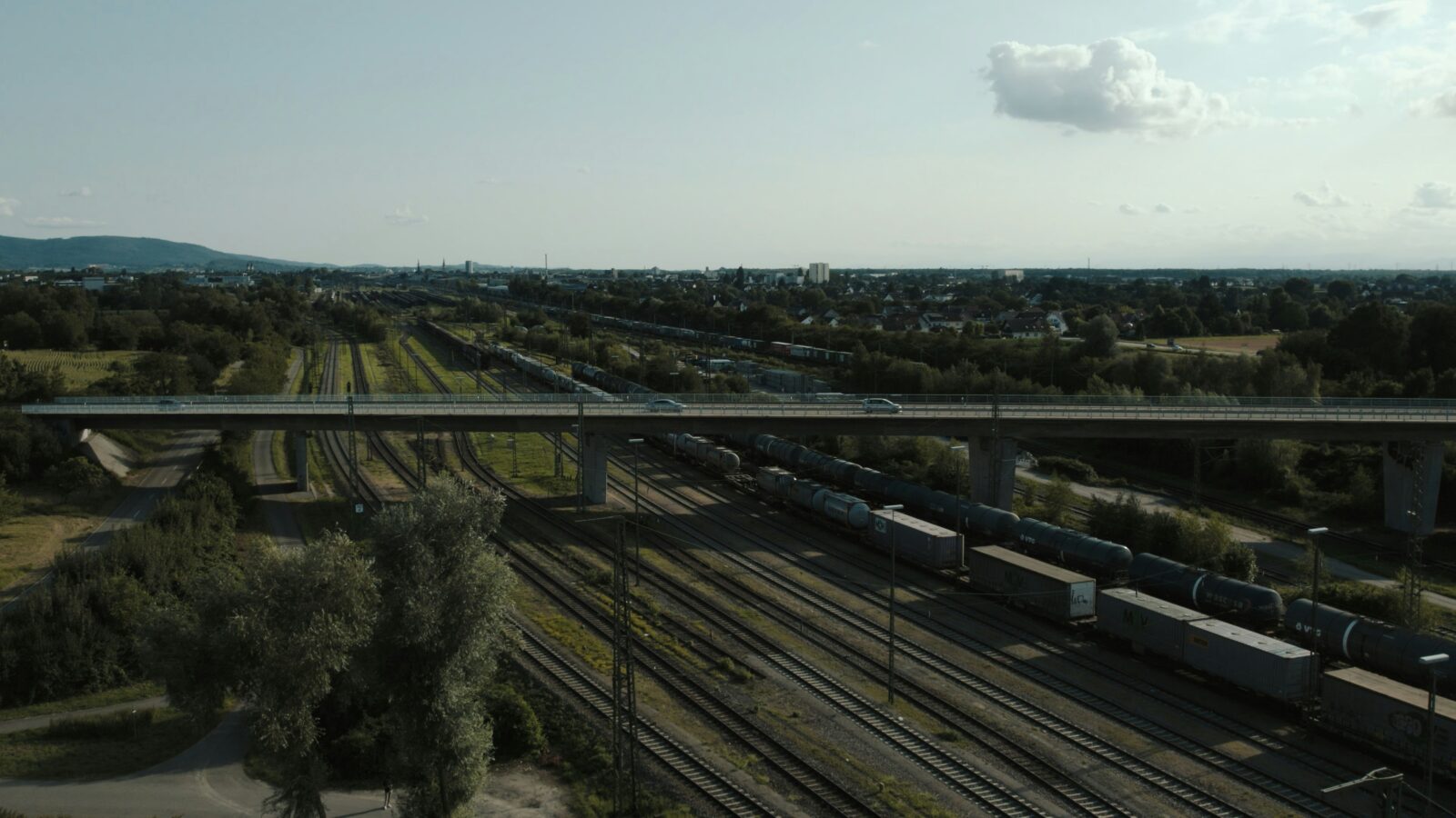 Flygfoto som visar en bro som korsar flera järnvägsspår med godståg, omgiven av grönska och en stad i bakgrunden under en molnig himmel.